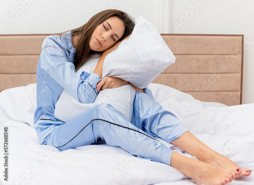 young beautiful woman in blue pajamas sitting on bed with pillow feeling positive emotions with closed eyes in bedroom interior on light background © HN Works