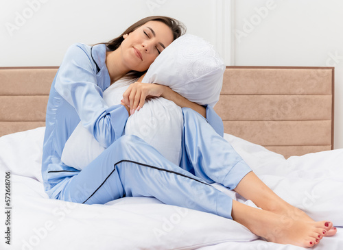 young beautiful woman in blue pajamas sitting on bed hugging pillow feeling positive emotions with closed eyes in bedroom interior on light background