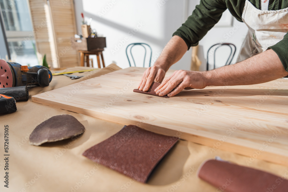 Cropped view of repairman sanding wooden board near sandpaper in workshop.