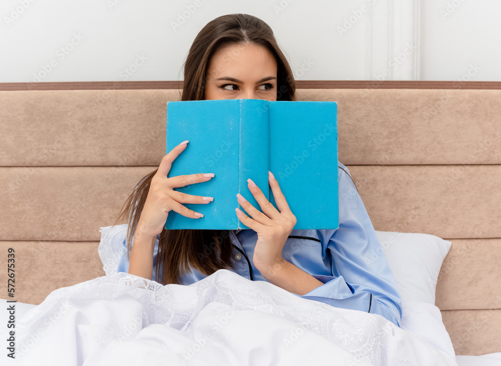 young beautiful woman in blue pajamas sitting on bed with book hiding face looking aside peeking over in bedroom interior on light background young beautiful woman in blue pajamas sitting on bed with book hiding face looking aside peeking over in bedroom interior on light background
