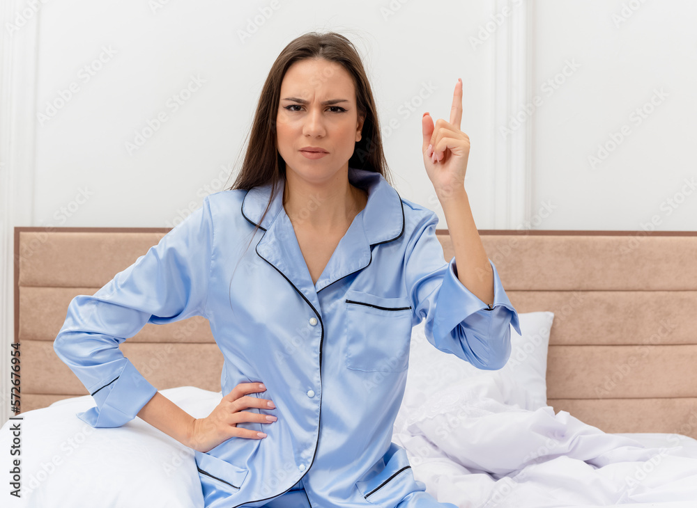 young beautiful woman in blue pajamas sitting on bed showing index finger with serious face in bedroom interior on light background young beautiful woman in blue pajamas sitting on bed showing index finger with serious face in bedroom interior on light background
