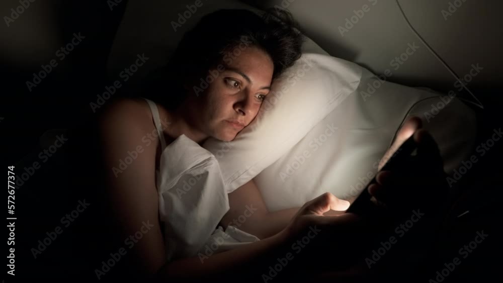 Woman laid in bed looking at smartphone screen with glowing light on face. Person using phone at night before sleep