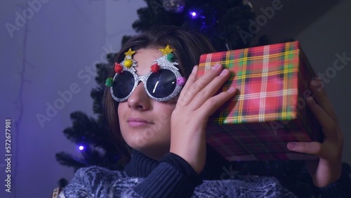 A teenager boy in funny New Year glasses shakes a box with a gift against the background of a Christmas tree and illumination