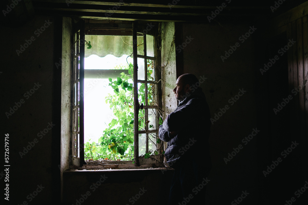 A man inside an old crumbling house near the window. Depression, loneliness, despair, decline, sadness concept.