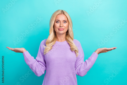 Photo of expert promoter middle aged woman wear purple knitted pullover hold palms empty space balance products isolated on cyan color background © deagreez