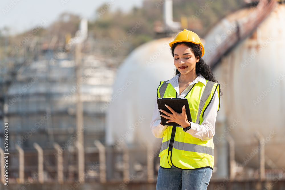 female engineer with hardhat with petrochemical factory background. asian woman holding tablet, plan and Walkie Talkie.