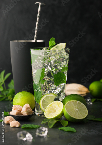 Glass of Mojito cocktail with ice cubes mint and lime on black board with spoon in shaker and wooden squeezer and fresh limes