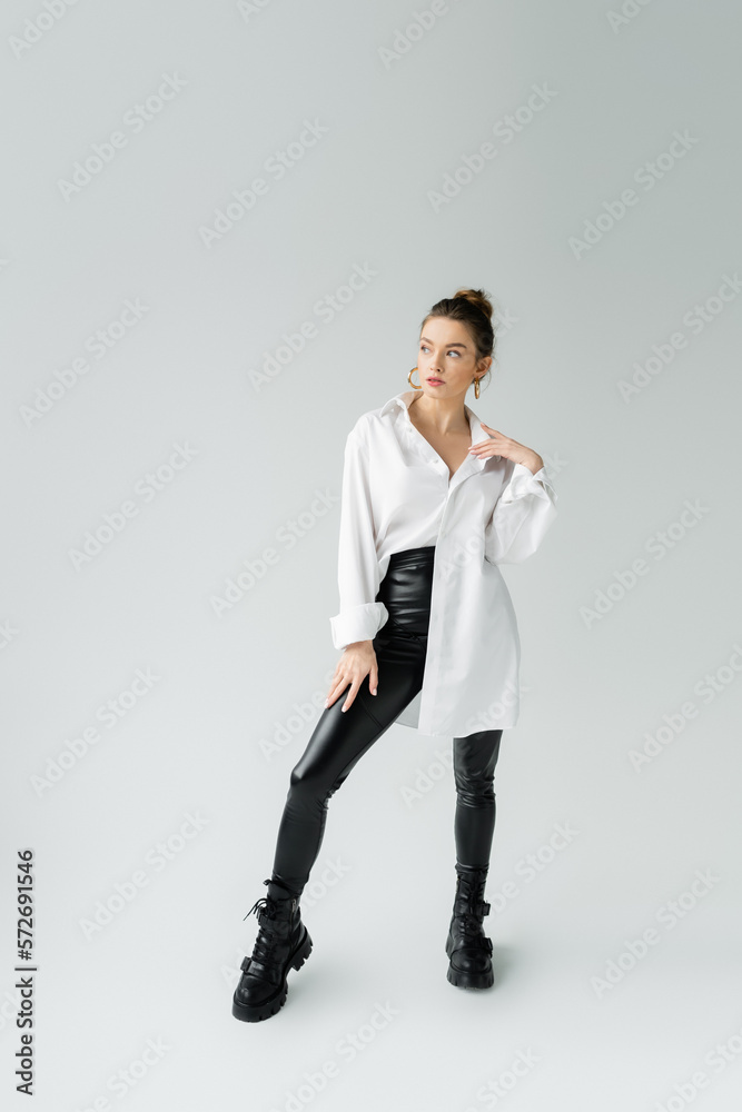 full length of trendy woman in oversize shirt and tight pants with rough boots looking away on grey background. full length of trendy woman in oversize shirt and tight pants with rough boots looking away on grey background.