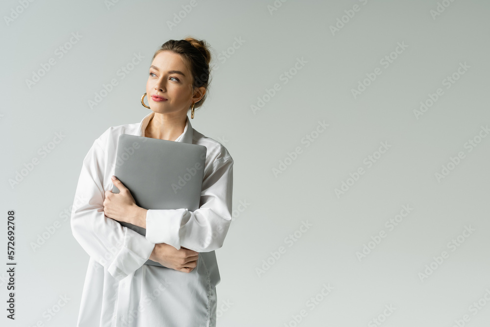 positive woman in white shirt holding laptop and looking away isolated on grey. positive woman in white shirt holding laptop and looking away isolated on grey.
