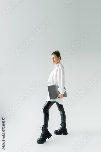 full length of stylish woman in black latex pants and rough boots posing with laptop on grey background.