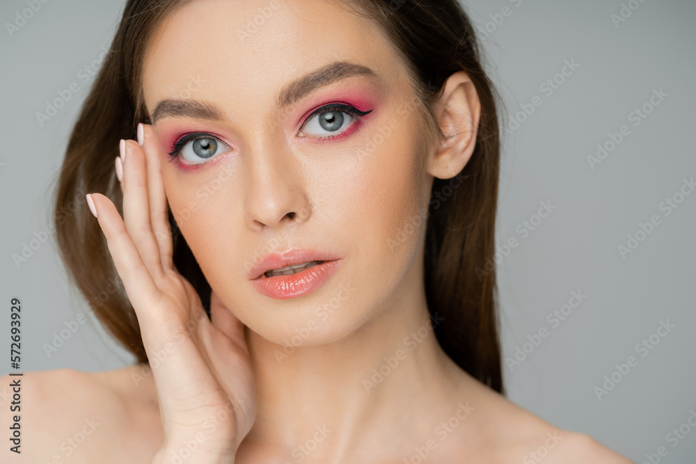 Portrait of young woman with pink makeup touching face isolated on grey.