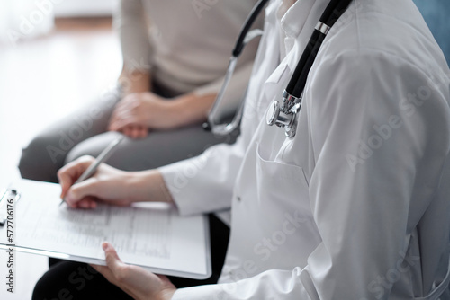 Doctor and patient sitting at sofa in clinic office. The focus is on female physician's hands filling up the medication history record form, close up. Medicine concept