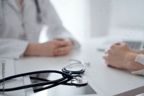 Fototapeta Naklejka Na Ścianę i Meble -  Doctor and patient sitting and discussing something at the desk in clinic. The focus is on the stethoscope lying on the table, close up. Medicine and healthcare concept