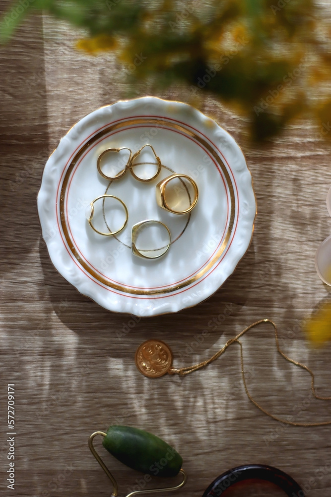 Retro bag, plate with gold rings and earrings, gold necklace, sunglasses, face roller, lit candles and mimosa flowers on wooden background. Flat lay. Retro bag, plate with gold rings and earrings, gold necklace, sunglasses, face roller, lit candles and mimosa flowers on wooden background. Flat lay.
