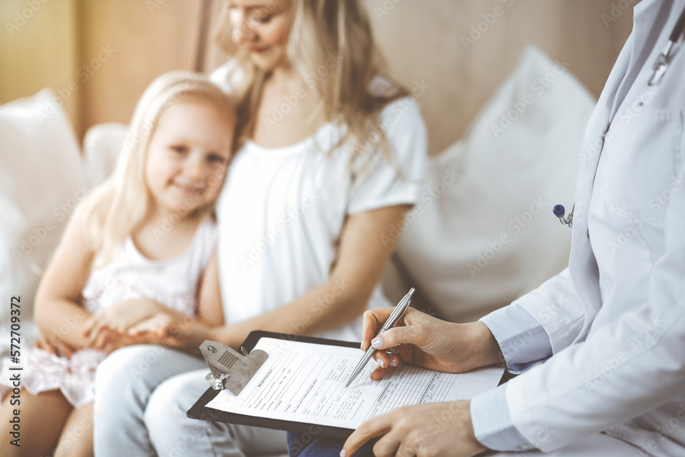 Doctor and patient. Pediatrician using clipboard while examining little girl with her mother at home. Happy cute caucasian child at medical exam. Medicine concept Doctor and patient. Pediatrician using clipboard while examining little girl with her mother at home. Happy cute caucasian child at medical exam. Medicine concept