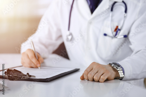 Unknown male doctor sitting and working with clipboard of medication history record in a darkened clinic  glare of light on the background  close-up of hands