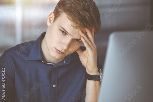 Young blond businessman feeling stress at workplace in a darkened office, glare of light on the background. Startup business means working hard and out of time