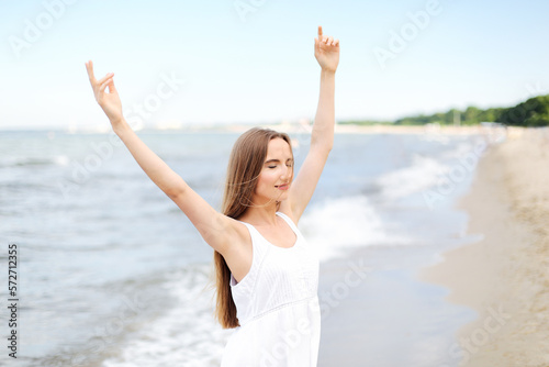Happy smiling woman in free happiness bliss on ocean beach standing with raising hands. Portrait of a multicultural female model in white summer dress enjoying nature  © rogerphoto