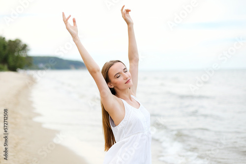 Happy smiling woman in free happiness bliss on ocean beach standing with raising hands. Portrait of a multicultural female model in white summer dress enjoying nature 