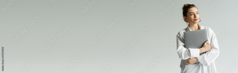 pleased woman in white shirt holding laptop while looking away isolated on grey, banner.