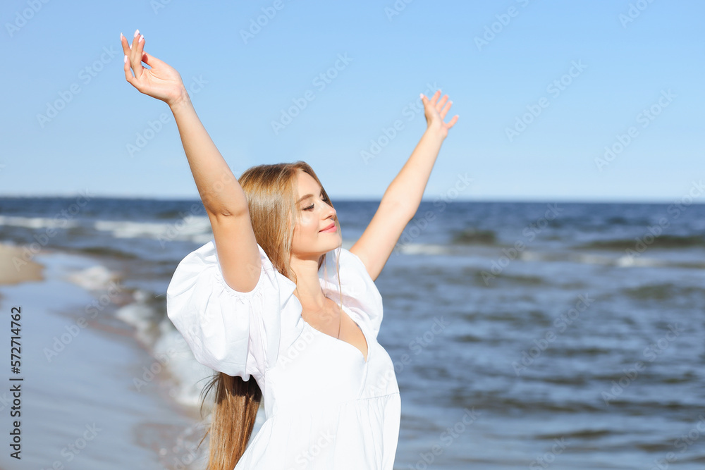 Happy, beautiful woman on the ocean beach standing in a white summer dress, raising hands Happy, beautiful woman on the ocean beach standing in a white summer dress, raising hands