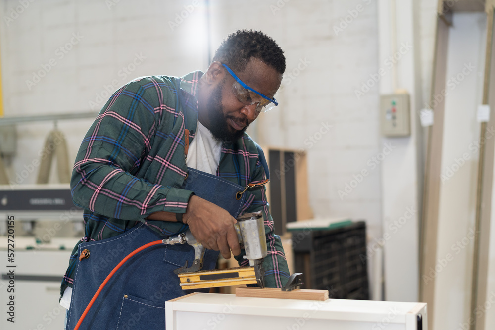 African American male carpenter using pneumatic nail gun shoot plank at wood workshop. Joiner male worker working with pneumatic staple gun making new furniture in wood factory African American male carpenter using pneumatic nail gun shoot plank at wood workshop. Joiner male worker working with pneumatic staple gun making new furniture in wood factory