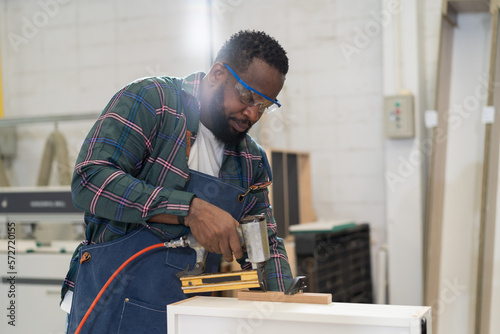African American male carpenter using pneumatic nail gun shoot plank at wood workshop. Joiner male worker working with pneumatic staple gun making new furniture in wood factory