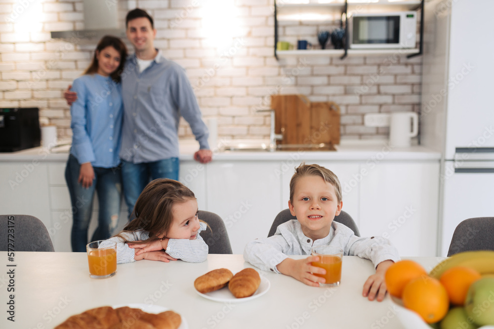 Happy family having breakfast on kitchen. Little girl with brother sitting on kitchen and smiling with their parents on background Happy family having breakfast on kitchen. Little girl with brother sitting on kitchen and smiling with their parents on background