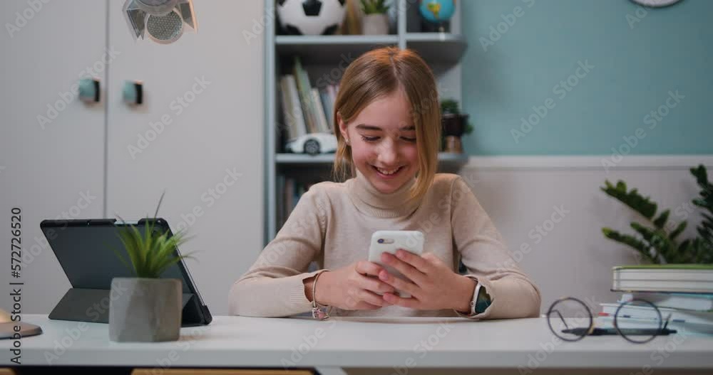 Good-looking smiling 10 years old girl holding mobile phone, using mobile apps, playing games near tablet device sitting at table in living room at home