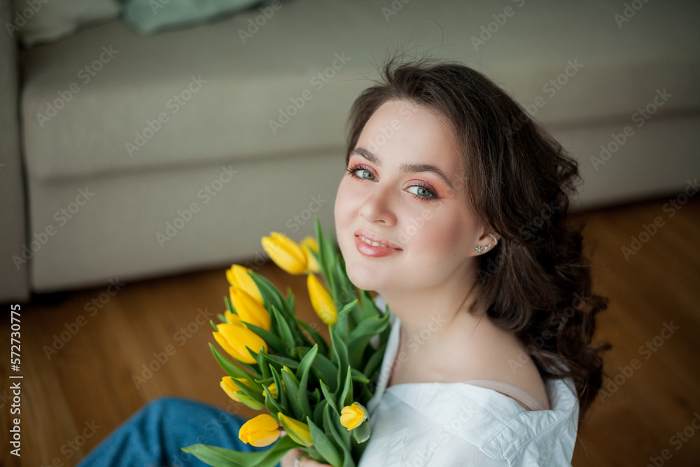 Spring portrait of young beautiful woman with blue eyes and curly hair with bouquet of yellow tulips in home interior. Happy model in jeans and white shirt. Mothers Day.