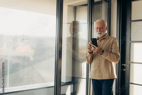 Handsome smiling senior man standing near panoramic window at home using smart phone technology. Cheerful senior citizen checking online banking mobile app. Older gray haired man typing on phone.