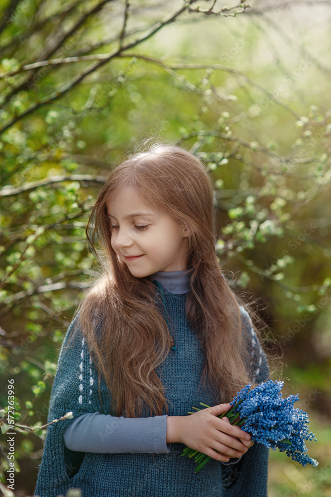 Pretty little happy smiling girl with long hair and bouquet of blue flowers in spring park with greenery and sunshine in background, cosy warm child outdoor portrait, tenderness and beauty of nature Pretty little happy smiling girl with long hair and bouquet of blue flowers in spring park with greenery and sunshine in background, cosy warm child outdoor portrait, tenderness and beauty of nature