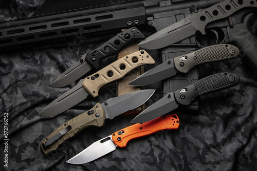A variety of pocket military knives lie near the m4 carbine. Gray camouflage back. photo