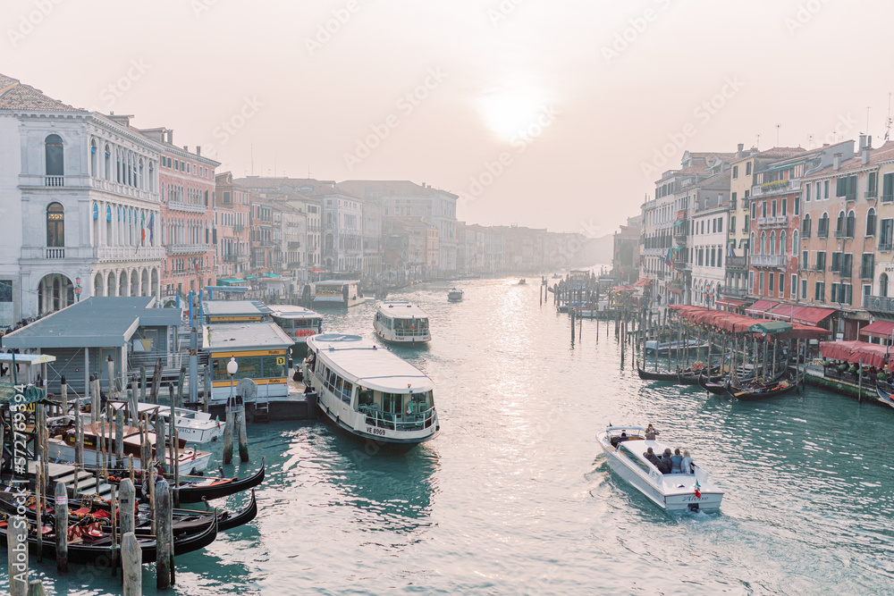 An impressive view over a river in Venice. The sky is overcast and gives a slight hint of the sun. Countless gondolas are docked at the harbors, waiting for their tourists.