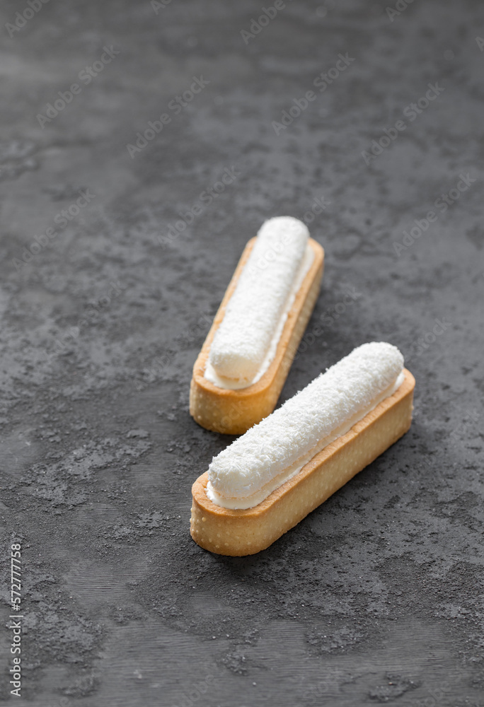 Two oval tartlets in the form of eclair with vanilla custard and delicate creamy jelly sprinkled with coconut chips on top. Dark gray background Two oval tartlets in the form of eclair with vanilla custard and delicate creamy jelly sprinkled with coconut chips on top. Dark gray background