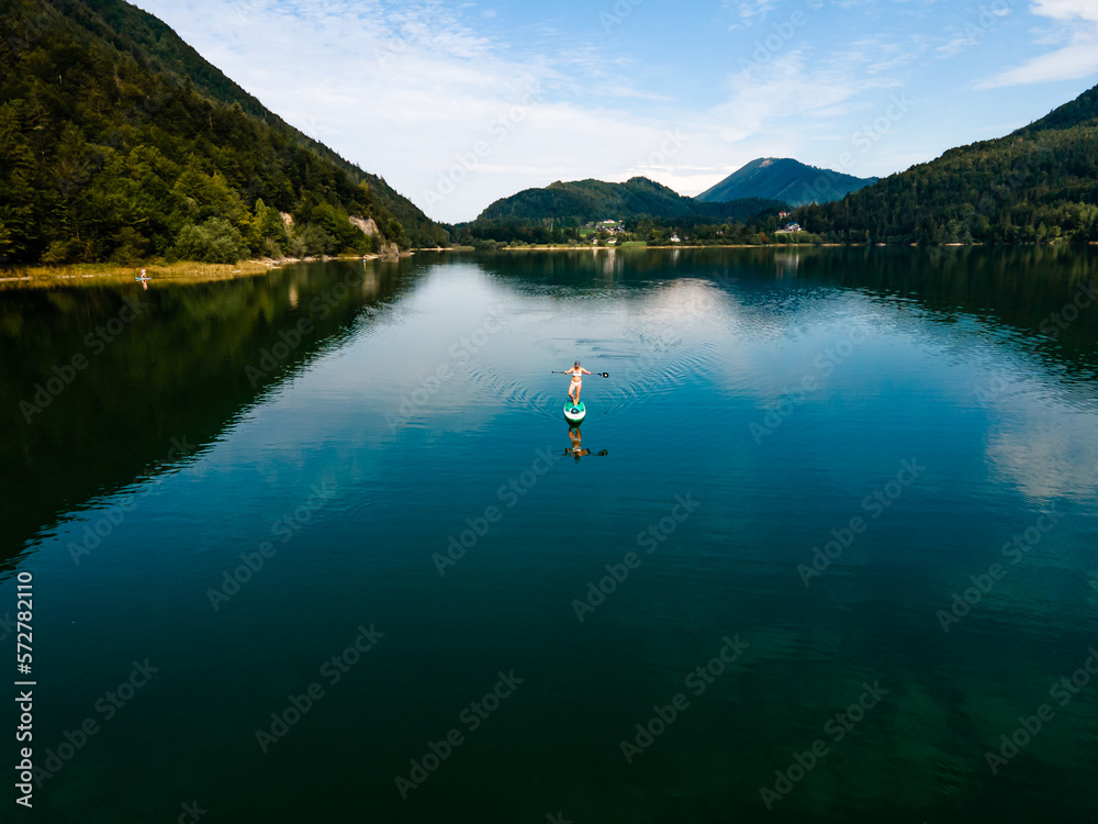 Woman Paddles a SUP Board on a Lake, Embraced by Majestic Mountains in background