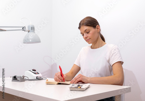 Beautiful nail beautician makes notes in notebook with pen, sitting at workstation, table in nail beauty treatment salon. Professional tools are on white table. Manicurist's routine. Horizontal plane photo