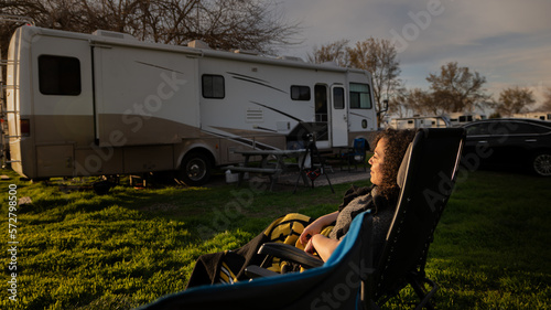 Young lady sitting in the sunlight of a sunset at campsite with Rv motorhome