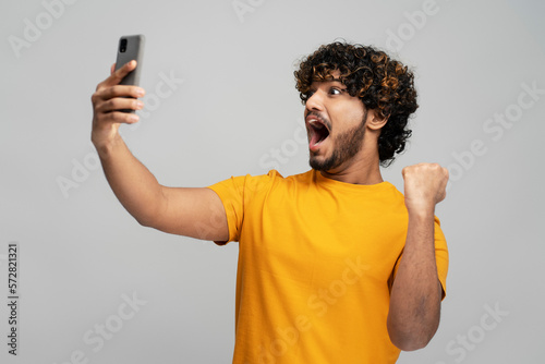 Overjoyed young curly bearded Indian man in yellow t-shirt holding a modern mobile phone in winning lottery, reading good news, isolated over gray background. Guy sports betting celebration success  photo