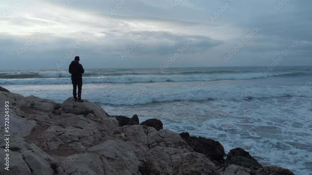 Epic drone shot gliding past a man standing on a cliff's edge while ferocious waves crash onto the shore Epic drone shot gliding past a man standing on a cliff's edge while ferocious waves crash onto the shore