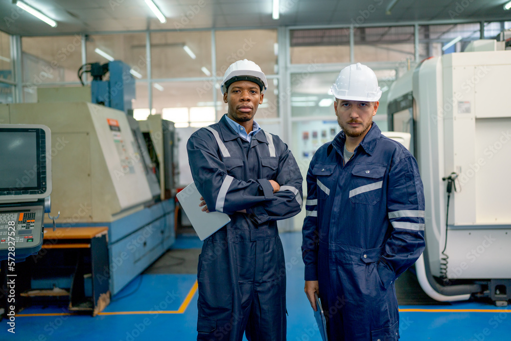 African American and Caucasian technician or engineer workers stand with arm crossed and look at camera also stay in front of factory machine in workplace.