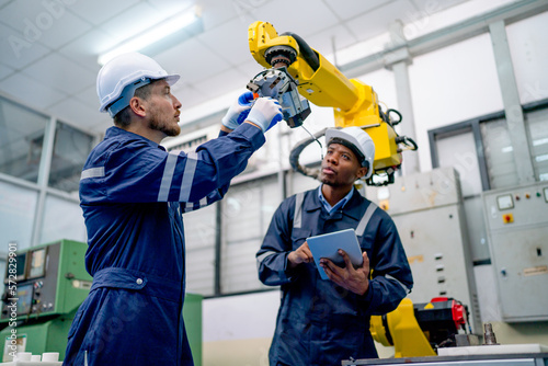 Caucasian technician hold and check part of robotic arm while his African American coworker hold tablet and record the data in workplace.