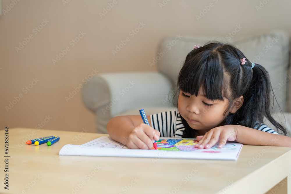 Front view medium to close-up portrait with copy space of cute 6-year-old pre-school Asian girl concentrating, enjoying drawing, doing homework in living room. Education, art, leisure concept. Front view medium to close-up portrait with copy space of cute 6-year-old pre-school Asian girl concentrating, enjoying drawing, doing homework in living room. Education, art, leisure concept.