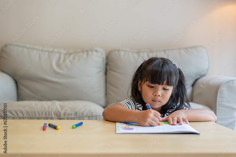 Front view medium shot portrait with copy space of cute 6-year-old pre-school Asian girl concentrating, enjoying drawing, doing homework in living room. Education, art, leisure, hobby concept. Front view medium shot portrait with copy space of cute 6-year-old pre-school Asian girl concentrating, enjoying drawing, doing homework in living room. Education, art, leisure, hobby concept.
