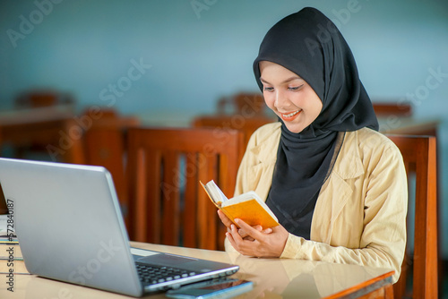 Beautiful young asian woman wearing hijab smiling and reading pocket Quran in between work, and there are books, laptop computer and mobile phone on her desk.