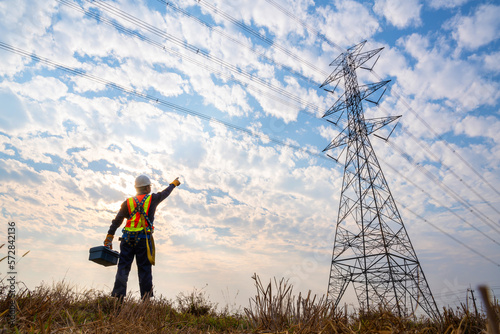A technician stands holding a toolbox while pointing at a high-voltage pole wearing an operator fall arrester with hooks for safety harnesses. maintenance goal concept. at high voltage pylons.