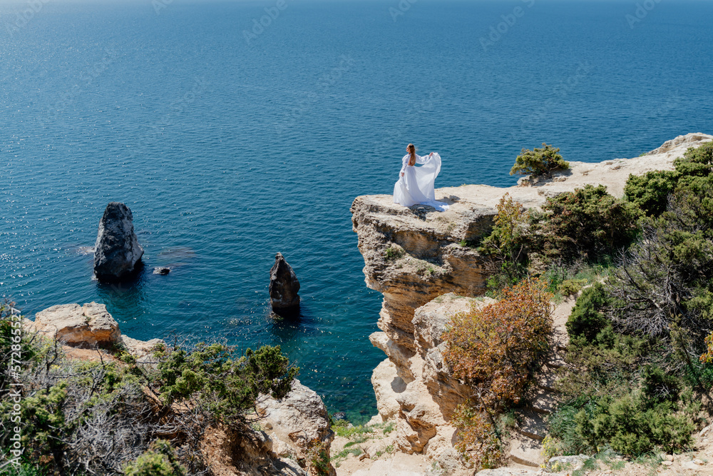 Woman in a white dress on the sea. Side view Young beautiful sensual woman in white long dress posing on a rock high above the sea at sunset. Girl in nature against the blue sky