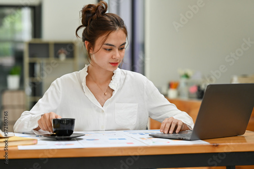 Charming young female employee holding coffee cup and checking business email on laptop computer.
