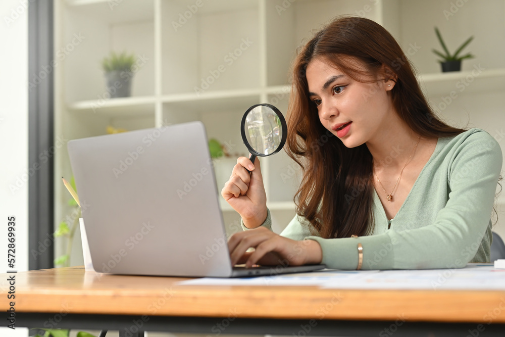 Beautiful young businesswoman hand holding magnifying glass and using laptop computer at office desk. Beautiful young businesswoman hand holding magnifying glass and using laptop computer at office desk.