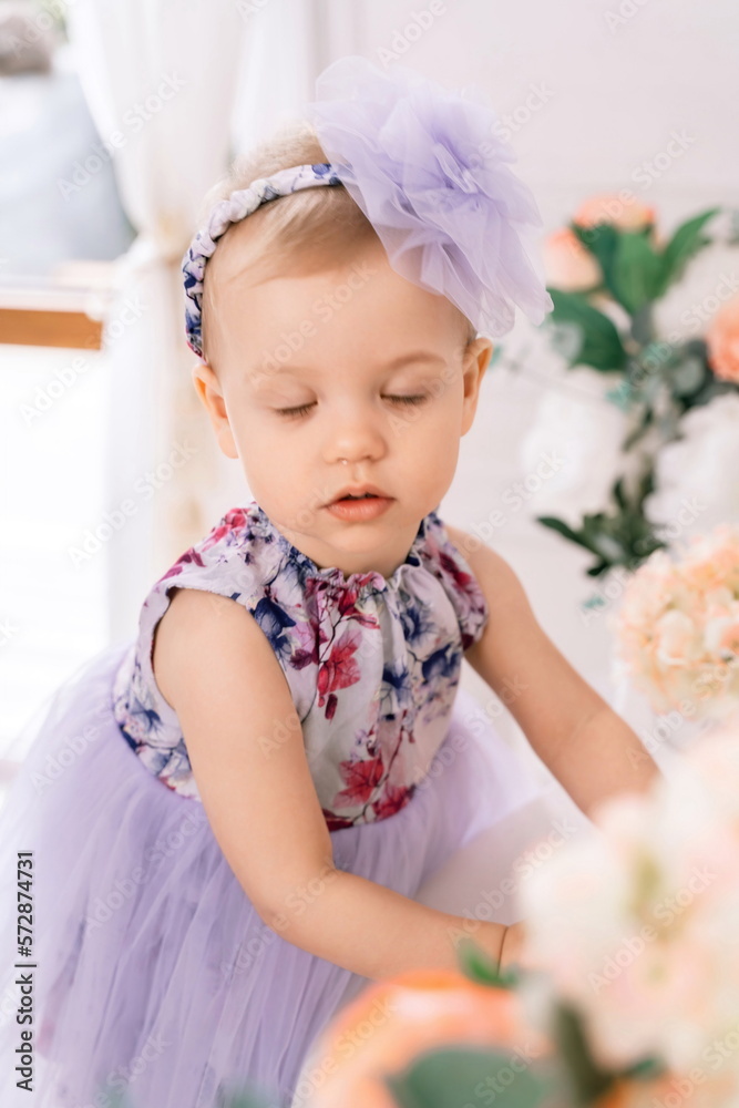 Baby girl elegant dress. A one-year-old girl in a puffy dress and a cute bow poses against the backdrop of a bright room with a dressing table and flowers. Baby girl elegant dress. A one-year-old girl in a puffy dress and a cute bow poses against the backdrop of a bright room with a dressing table and flowers.